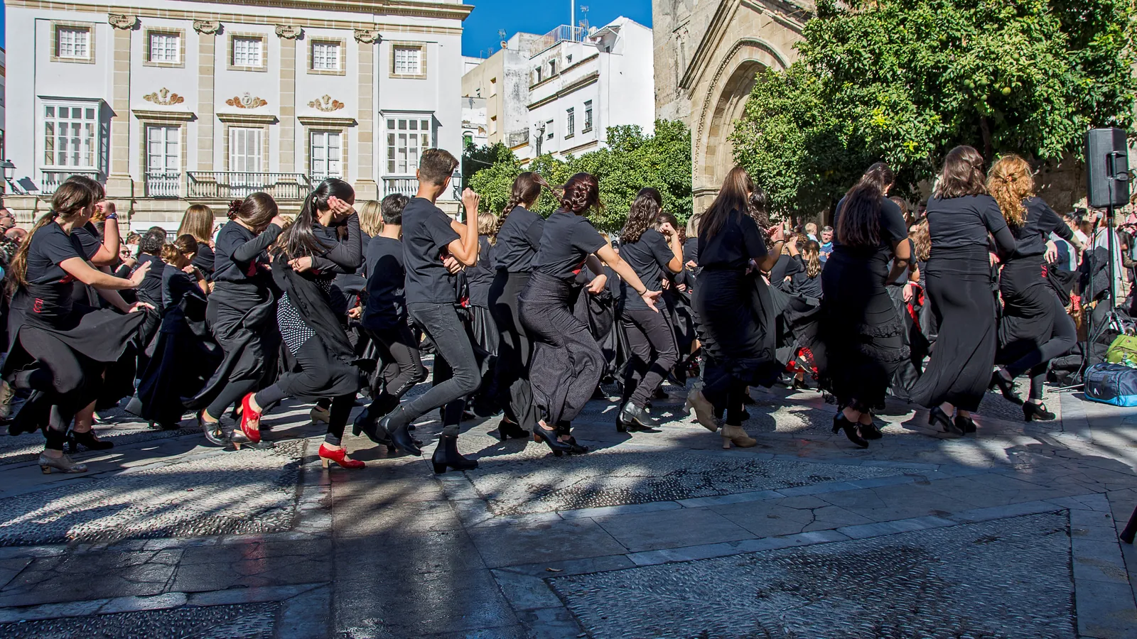 Dancers in black skirts and tops dance together in a sunlit square in Jerez, beneath a white town house and a stone church doorway