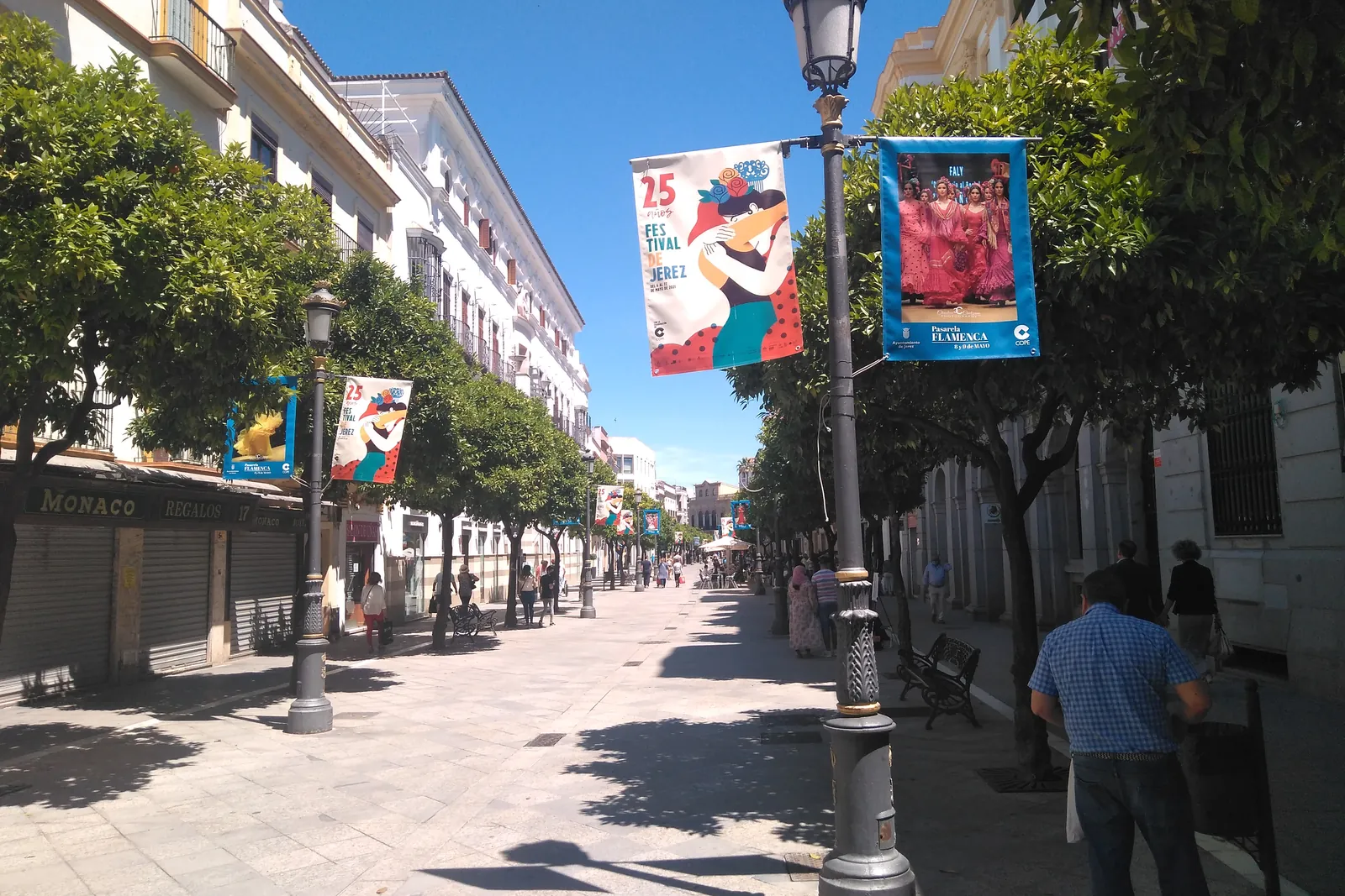 Colourful flamenco festival banners hung from lampposts along a pedestrian street in central Jerez, under orange trees