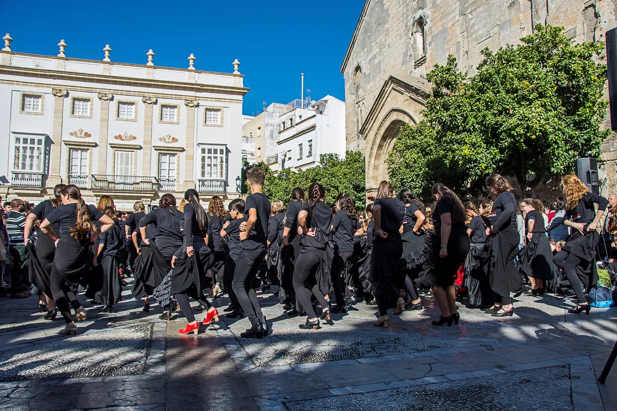 A large group of dancers in black skirts and tops performing together in a sunlit square in Jerez, with a white town house on the left and a stone church doorway on the right
