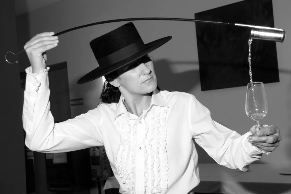 A woman in a wide-brimmed black hat and a ruffled white shirt pours sherry from a long-handled cup held high into a glass in her other hand, in black and white